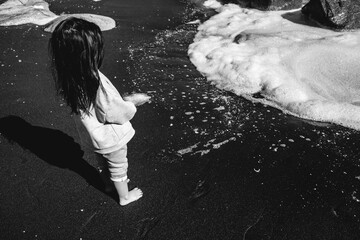 Little girl barefoot in the sand in front of the sea with foamy waves (in black and white)