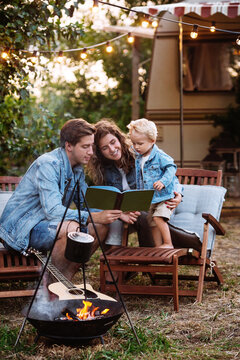 Recreation at campfire with little kid: young parents read book to small son on vacation trip in caravan car. Hipster family travel by camper trailer with child together. Summertime nature relaxation
