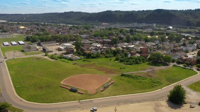 A slowly orbiting aerial view of a small Pennsylvania river town. A baseball sandlot field in the foreground and the Ambridge Bridge over the Ohio River in the distance.  	