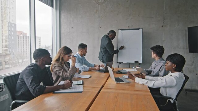 Multiethnic group businesspeople diverse managers workers multiracial business team sitting at office table meeting brainstorming listening african mature boss leader standing writing on white board