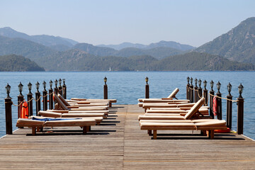 Beach vacation on sea resort, lounge chairs on a wooden pier against a misty mountains. Empty sun beds on green island background