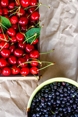 Cropped bowl of blueberries and box, crape of red sweet cherries with tail on craft wrinkled old paper background. Vertical. Organic food. Top view. Summer fruits. Flatly. Still life