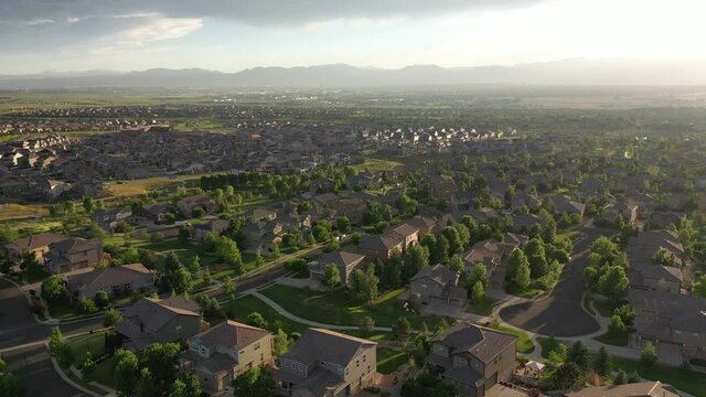 Aerial drone view of sprawling suburban housing development surrounded by green trees and parks in late evening with mountains in distance