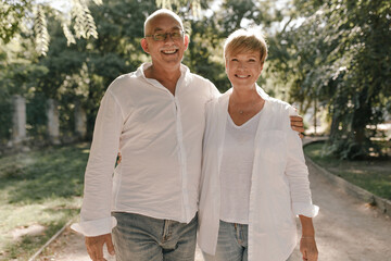 Old man with grey mustache and eyeglasses in white stylish shirt and jeans hugging her smiling wife with blonde hair in light blouse in park..