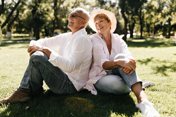 Smiling man with grey hair and mustache in glasses, long sleeve shirt and jeans sitting on grass with blonde lady in hat and striped blouse in park..