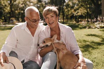 Cheerful lady with short blonde hair in pink shirt laughing, hugging dog and sitting on grass with...