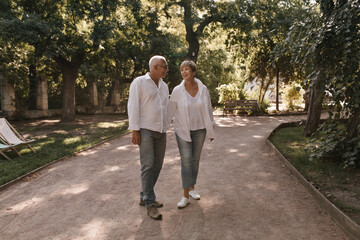 Fototapeta premium Full length photo of happy man with grey hair in white shirt and jeans smiling and posing with blonde lady in light blouse in park..