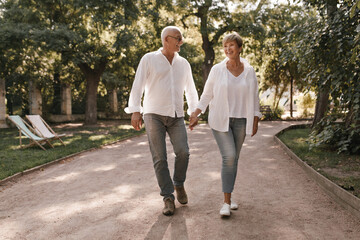 Fashionable lady with short hair in white blouse, jeans and sneakers smiling, walking and holding hands old man with glasses in shirt outdoor..