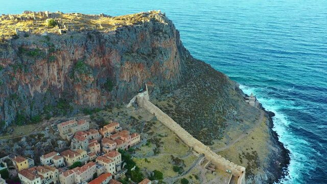 Aerial of the small ancient village Monemvasia in Greece. The village is settled on a hillside, facing the Mediterranean ocean. The drone hovers over the village and ocean, and is slowly coming down. 