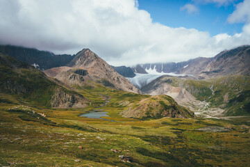 Fototapeta premium Scenic alpine landscape with mountain lake in sunlit green valley and glacier under cloudy sky. Awesome sunny scenery with beautiful glacial lake in sunlight against great mountain range in low clouds