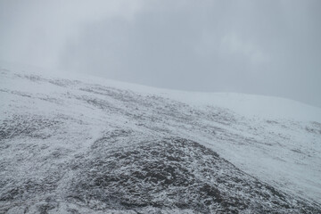 Foggy mountain landscape with white snow on black rocks in cloudy sky. Misty mountain minimalism of snowbound mountain top in low clouds. Minimalist nature background of snowy mountain peak in fog.