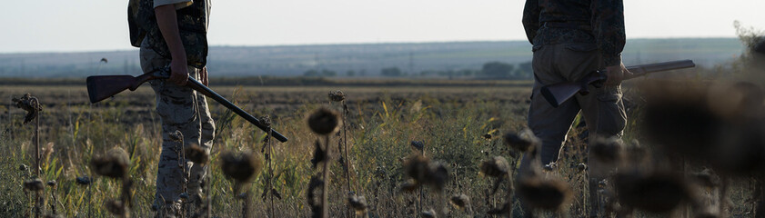 Duck hunters with shotgun walking through a meadow.