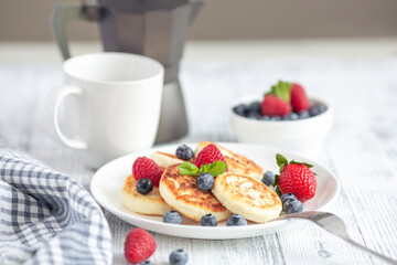 Cottage cheese pancakes with fresh berries, cup of coffee and coffee maker on white wooden table. Tasty breakfast food. Syrniki