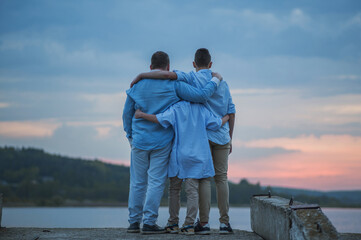 father with two sons in blue shirts hug and look at the view against the sunset background, back view, general plan