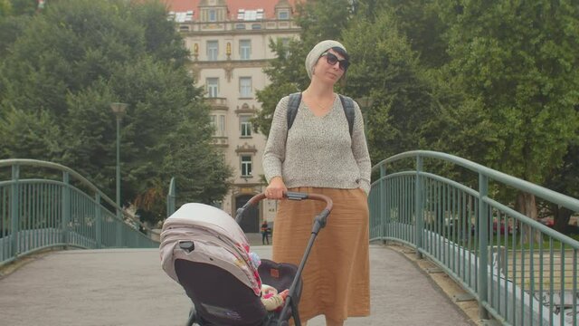 A young mother in a dress holds a stroller with her hand on the bridge. Glasses are on. The easy mood of the parent during the walk. A smile on your face.