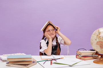 Young lovely school girl with pigtails and freckles in clear glasses looking away and holding book on her head on isolated background..