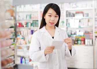Fototapeta na wymiar Chinese female looking medicine with notebook near shelves in pharmacy. High quality photo