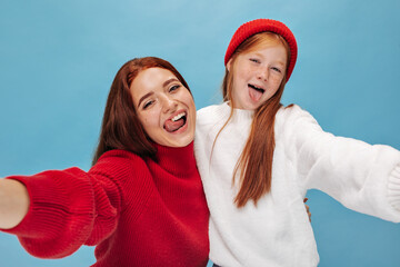 Stylish young girl with ginger hair and cute freckles in red cap shoes tongues and makes photo with bright sister in modern sweater..