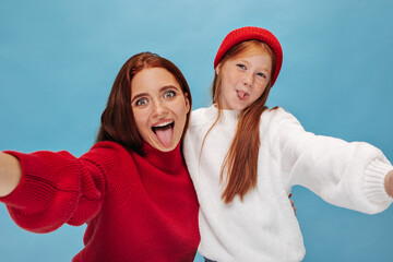 Funny cheerful two women in wide sweater showing tongues and takes selfie on isolated background. Lovely sisters in red outfit looking into camera..