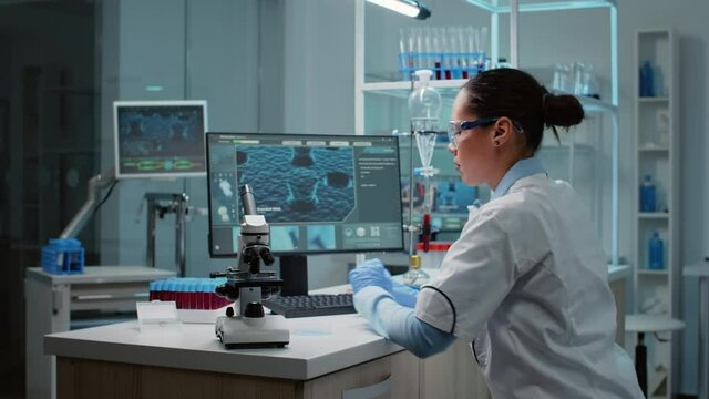 Biochemistry specialist doing research in laboratory with microscope, petri dish, vacutainers and analyzing dna animation on computer. Scientific woman looking at bacteria sample on plate