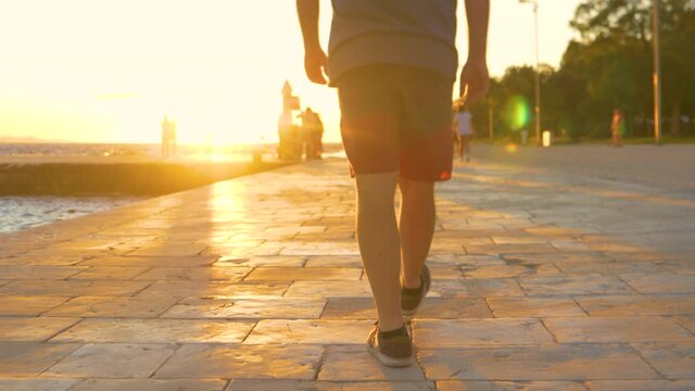 LOW ANGLE, CLOSE UP, LENS FLARE, DOF: Unrecognizable male tourist in shorts and sneakers walks along the picturesque Zadar promenade on a sunny summer evening. Man strolls along the Croatian coast.