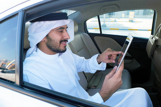 Emirati Arab using electronic tablet device  wearing traditional Dishdasha Kandoora while inside the car