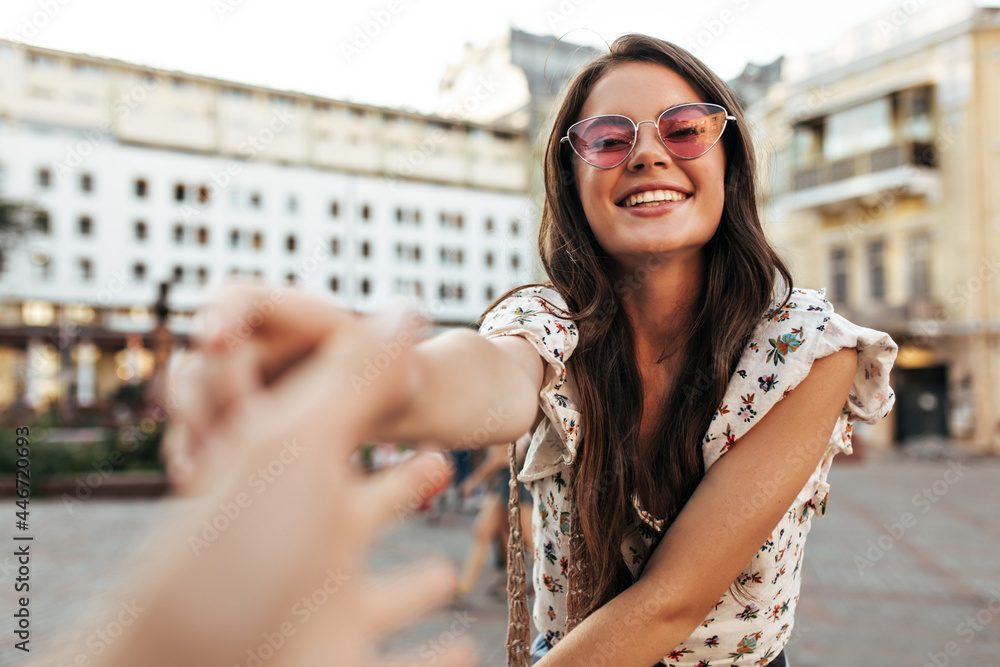 Sticker Tanned brunette curly woman in pink sunglasses reaches to someones hand. Attractive girl in stylish blouse smiles sincerely outside.