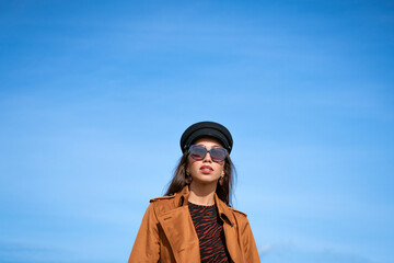Female portrait of a beautiful young woman of Caucasian appearance in a black cap against a blue sky in sunglasses and a jacket