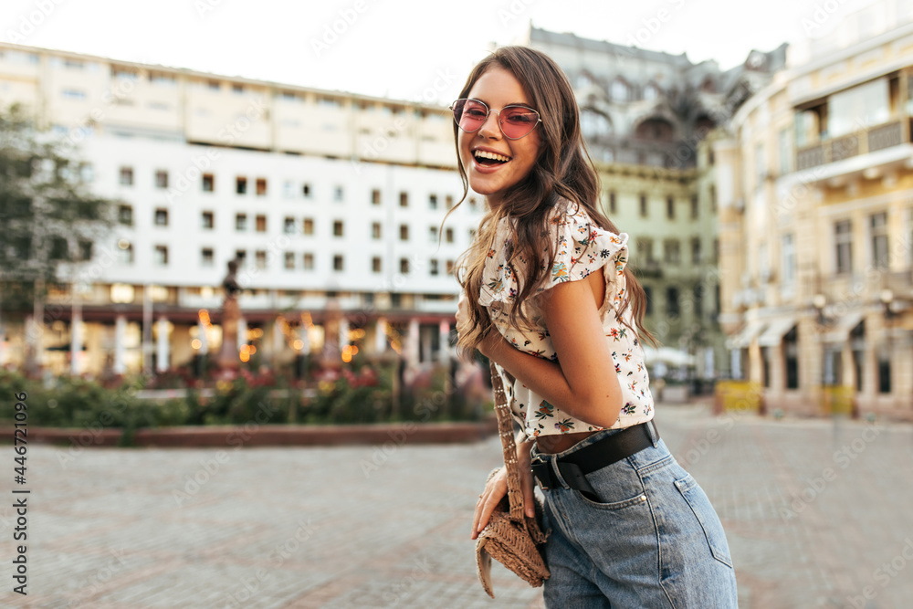 Sticker Cheerful brunette curly woman in pink sunglasses, denim pants and stylish floral top laughs and poses on great mood outdoors.