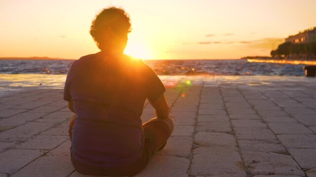 SLOW MOTION, LOW ANGLE, LENS FLARE: Unrecognizable young man sits on the stone ground of a scenic promenade in Zadar and observes the sea at golden sunset. Male tourist enjoys the summer evening view.