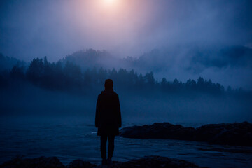 Dark human silhouette in a thick fog against the background of forest, hills and mountain river....
