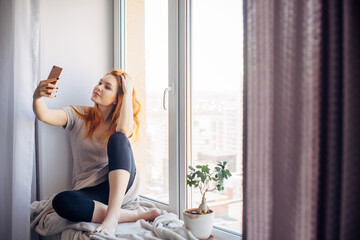 Pretty young woman with long red hair takes a selfie on her smartphone while sitting on the windowsill. Beautiful girl with a phone in a home environment. Technology and social networks concept.