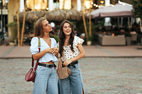 Charming young girls in florals colorful blouses and stylish denim pants walk in city and smile sincerely outdoors.
