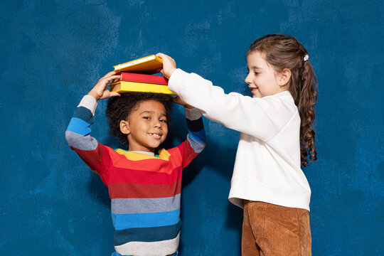 Two smiling diverse schoolchildren with textbooks, caucasian schoolgirl putting pile of books on head of mixed-race schoolboy on blue background. Back to school, children and education concept - Powered by Adobe