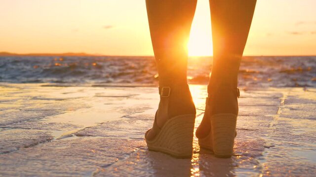 SLOW MOTION, LOW ANGLE, CLOSE UP, COPY SPACE, LENS FLARE, DOF: Unrecognizable female tourist in high heels observes the sunset from stone walkway running along the scenic shoreline of Zadar, Croatia.