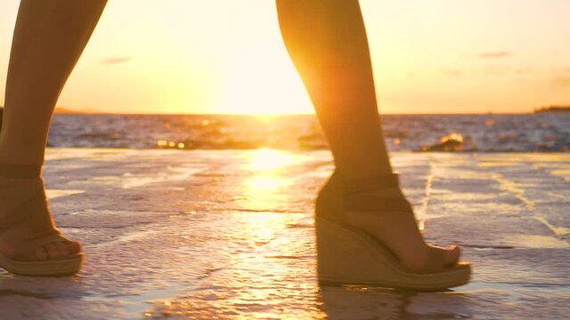 SLOW MOTION, LOW ANGLE, LENS FLARE, DOF: Female tourist goes for a stroll along the idyllic shore of Zadar, Croatia at golden sunset. Young woman in high heels walks along the scenic stone promenade.
