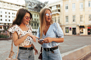 Brunette curly girl in white top and pink sunglasses smiles and points at phone screen of her friend. Blonde woman in blue blouse looks away outdoors.