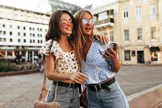Attractive tanned girls in loose stylish jeans and floral blouses smile and hug. Blonde and brunette women pose outdoors. Charming lady holds phone.