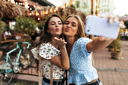 Pretty young attractive girls blow kiss and take selfie in good mood outside. Cheerful woman in blue blouse holds cellphone.
