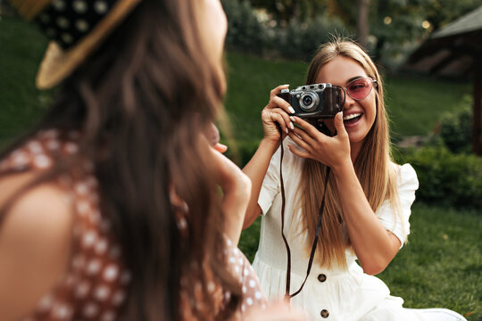 Charming blonde woman in stylish white dress and red sunglasses smiles and takes photo of her friend. Tanned girl holds retro camera. Brunette lady in boater has picnic. Blurred effect.