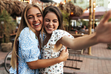 Joyful brunette and blonde women hug and sincerely smile outside. Beautiful lady in white floral blouse takes selfie with girlfriend at street.