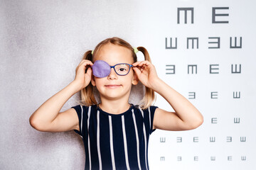 Happy little girl wearing glasses and eye patch or occluder with blurry eye test chart in the...