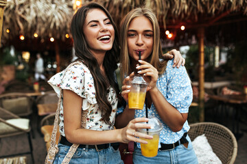 Excited happy brunette curly woman and cheerful blonde tanned girl in floral blouses drink lemonade and smiles outside.