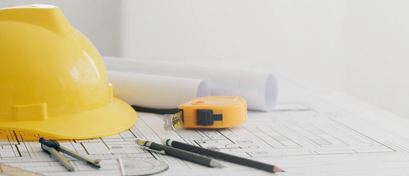 Yellow hard safety helmet hat and the blueprint, pen, ruler, protractor, and tape measure on the table at the construction site.for safety project of workman as engineer or worker.banner copy space.