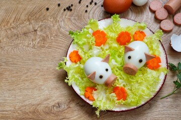  Boiled egg pigs,cute piglets made from boiled eggs,sliced sausage and black peppers on plate with green salad and flower shaped carrots with wooden table.Art food idea for kids.Top view.Copy space © KANJANA