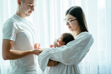 Fototapeta premium Emotional family lovely moment. Young cheerful parents wearing white clothes holding their little newborn baby girl, looking at her and smiling. Backlight with window on background.