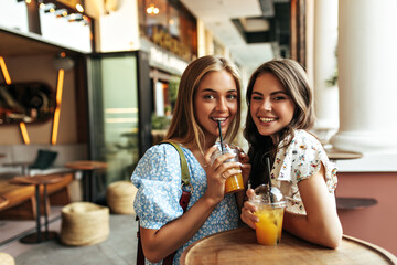 Good-humored young blonde and brunette women in trendy floral blouses smile sincerely, drink lemonade and rest in street restaurant.