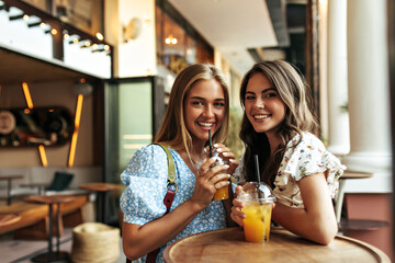 Happy cheerful brunette and young attractive blonde girls in stylish floral blouses drink sweet lemonade and smile sincerely in street cafe.