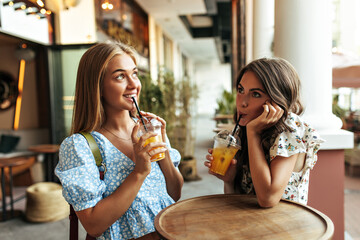 Cure young brunette and blonde happy women in stylish cropped floral blouses communicate, drink sweet lemonade and sit in cozy street cafe.