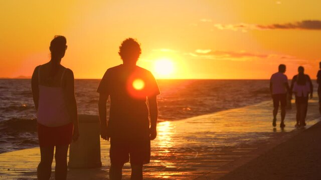 SLOW MOTION, SILHOUETTE, LENS FLARE, DOF: Young woman and man walk along a scenic coastal promenade at sunset. Unrecognizable male and female tourist go for a stroll during their summer vacation.